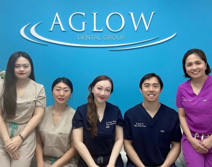 Aglow Group photo of five dental professionals from Aglow Dental Group standing in front of a blue wall with the Aglow Dental Group logo. Four women and one man, all in different colored scrubs, smile at the camera. The two women on the left wear beige scrubs, the central woman and the man wear navy scrubs with embroidered names, and the woman on the right wears purple scrubs.